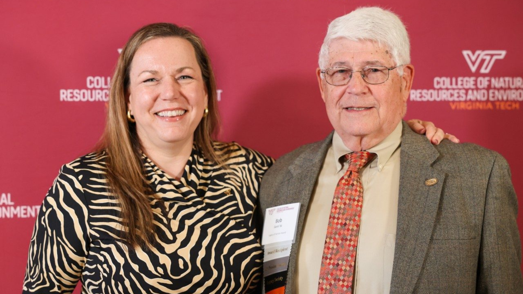 This photo shows two people standing side by side in front of a red backdrop, smiling at the camera. The backdrop features branding for Virginia Tech’s College of Natural Resources and Environment.